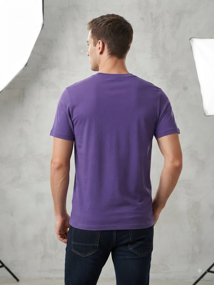 A man wearing a purple short-sleeve crewneck t-shirt, shown from the back, paired with dark blue jeans in a studio setting with a textured gray wall and softbox lights.
