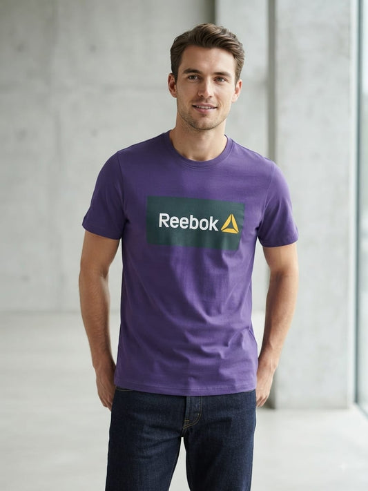 A man wearing a purple short-sleeve crewneck t-shirt and dark gray pants, photographed from the back in a bright, minimalist indoor setting.