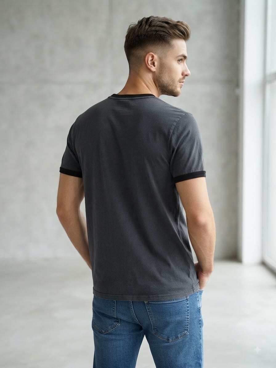 Man wearing a dark gray graphic t-shirt with a classic red and white logo box on the chest, paired with blue jeans and white sneakers, standing in a minimalist studio with a concrete-textured wall.