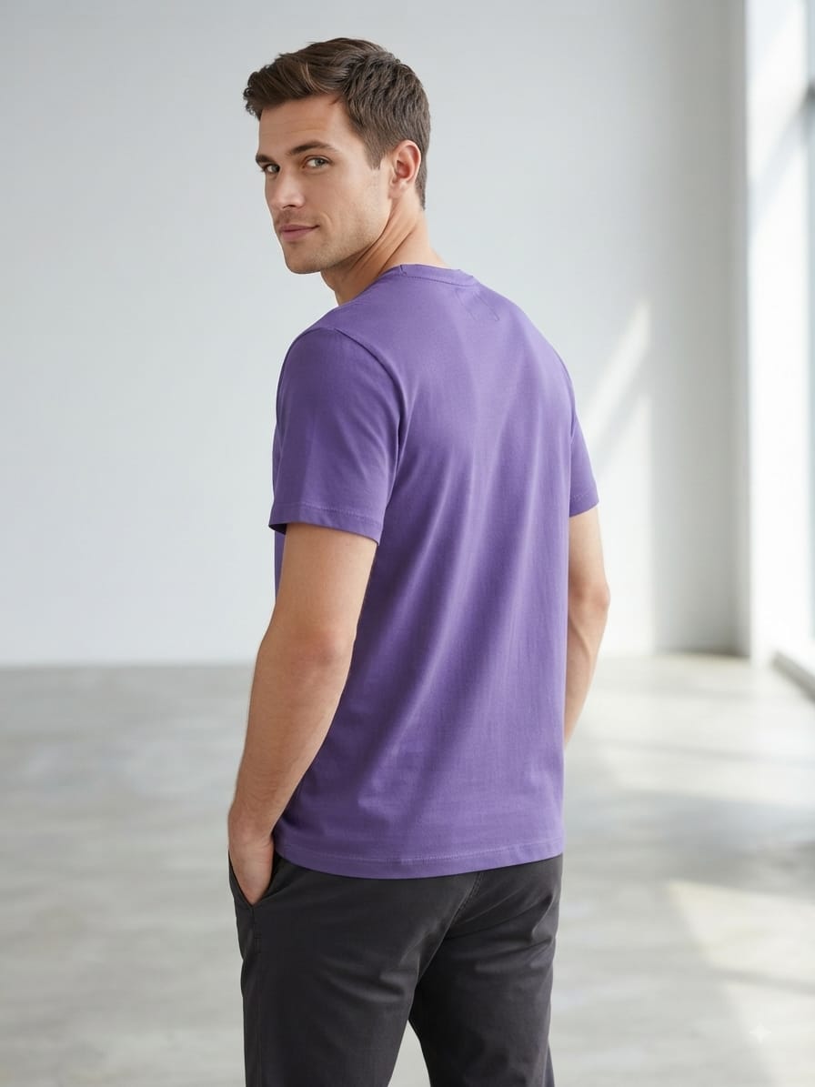 A man wearing a purple short-sleeve crewneck t-shirt and dark gray pants, photographed from the back in a bright, minimalist indoor setting.