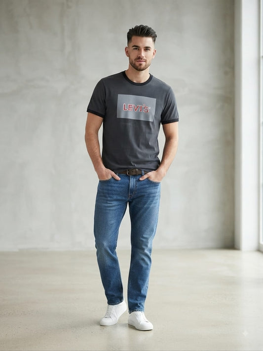 Man wearing a dark gray graphic t-shirt with a classic red and white logo box on the chest, paired with blue jeans and white sneakers, standing in a minimalist studio with a concrete-textured wall.